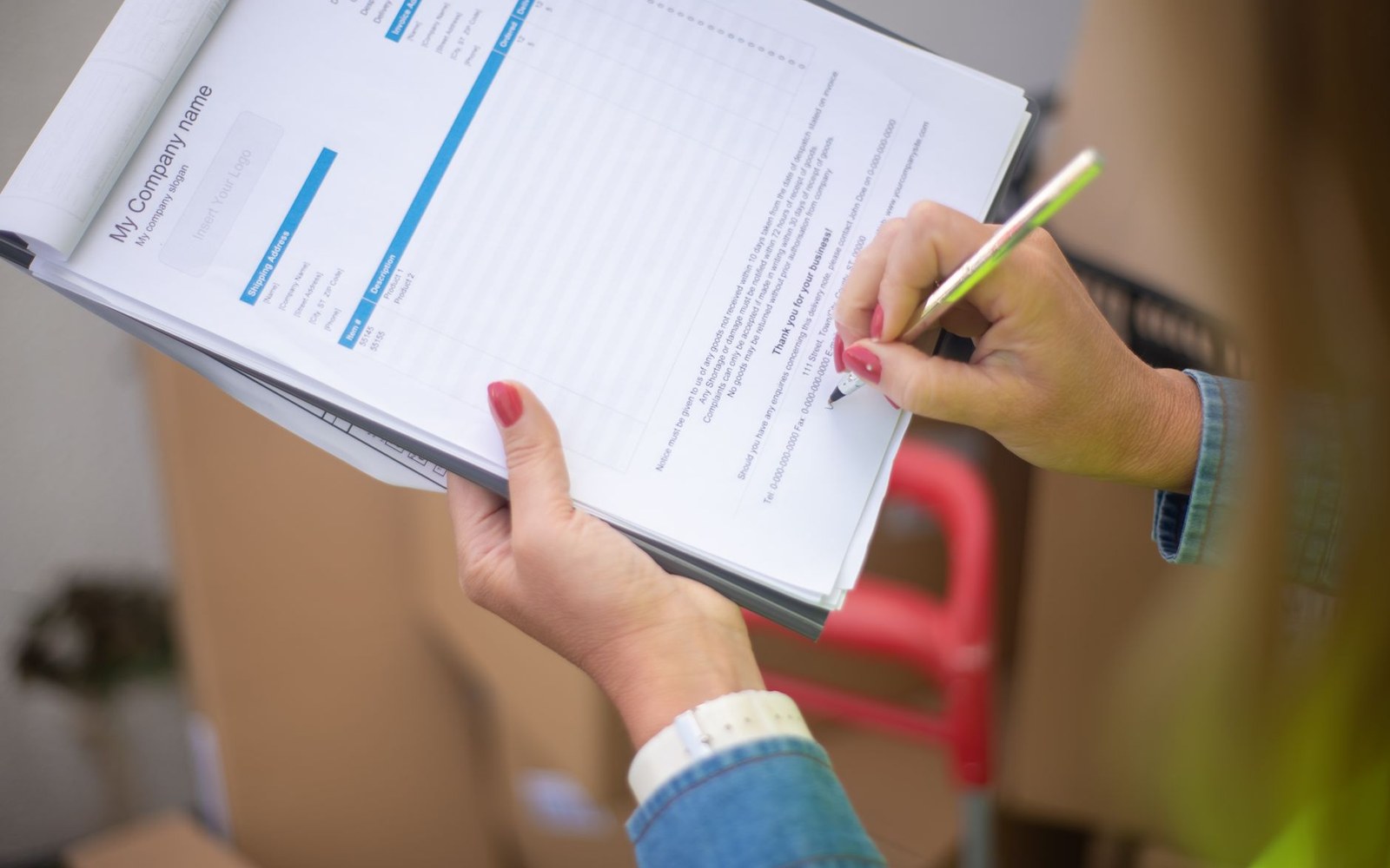 Hands writing on a company invoice form with a pen amid stacked cardboard boxes
