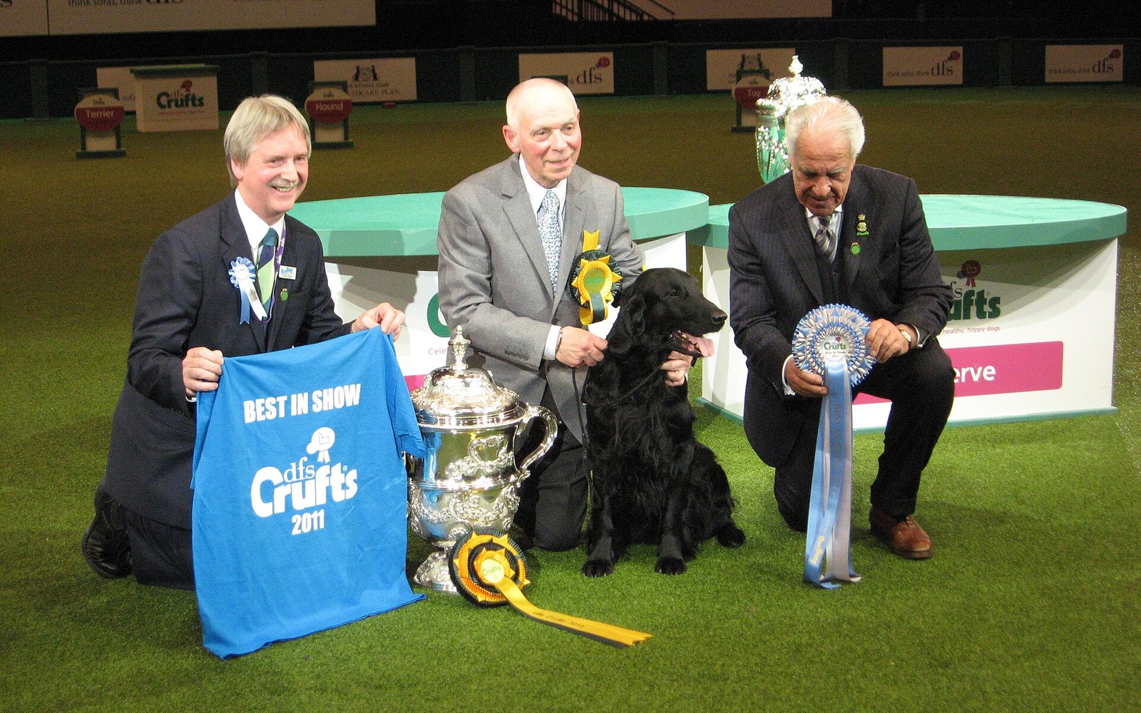 Three men kneel with a black flat-coated retriever and Best in Show trophy at Crufts 2011