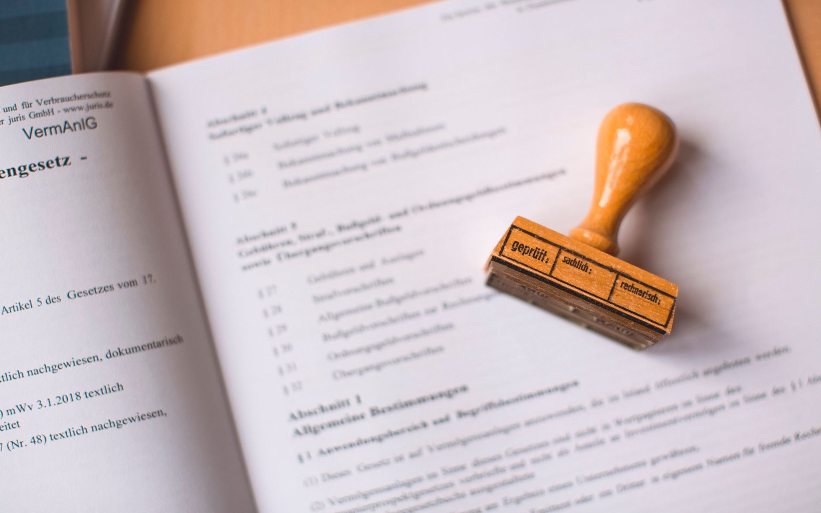 A wooden approval stamp reading "geprüft sachlich rechnerisch" resting on an open German legal text
