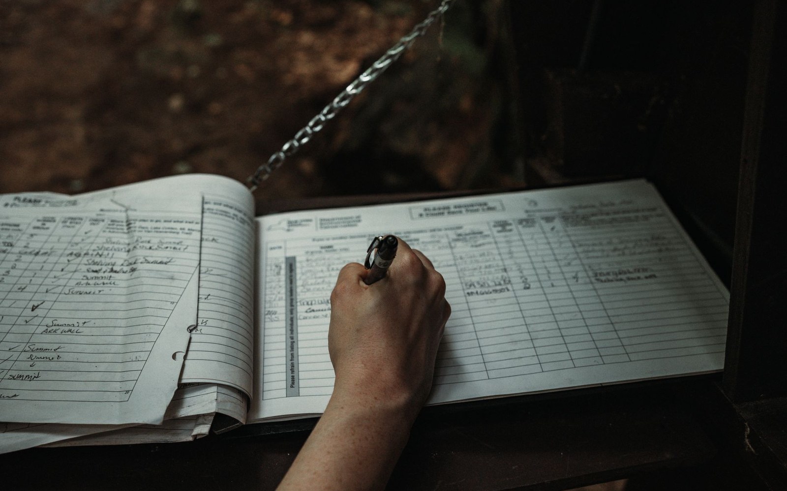 A hand writes in a ruled logbook attached to a chain outdoors