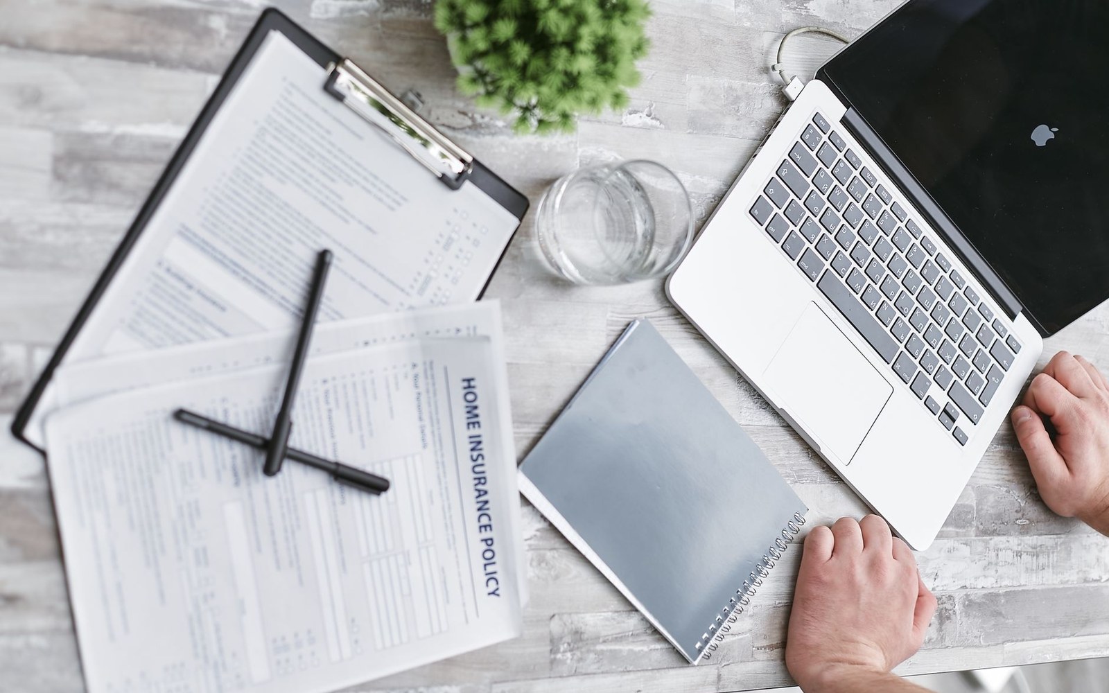 Hands typing on a laptop beside a home insurance policy document and notebook on a desk